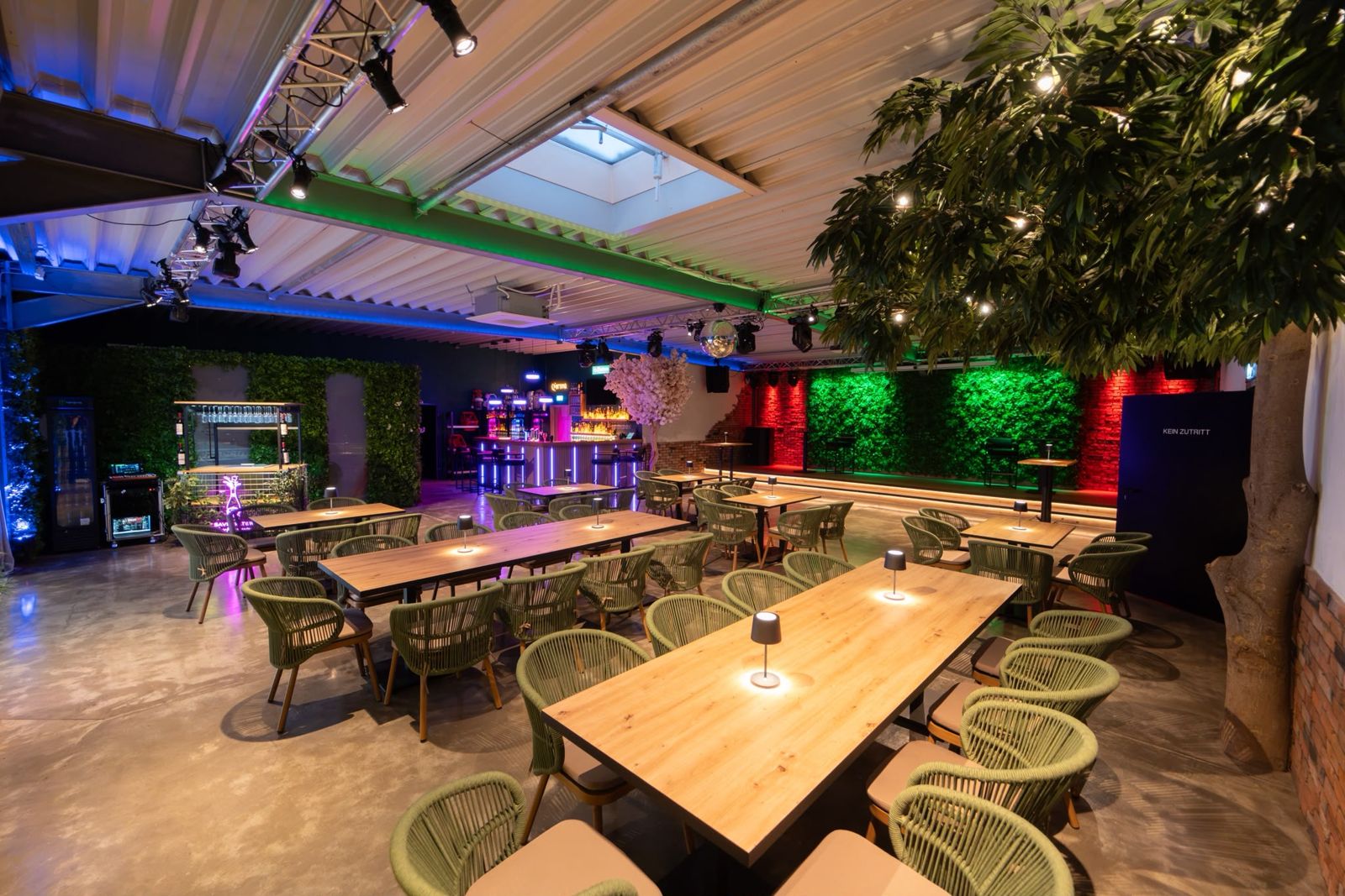 Saal mit langen Holztafeln, Rattanstühlen und begrüntem Baum